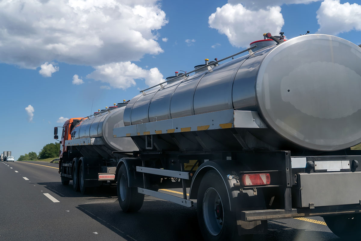 Stainless steel tanker truck pulling two tank trailers along a divided highway under a blue sky with white clouds