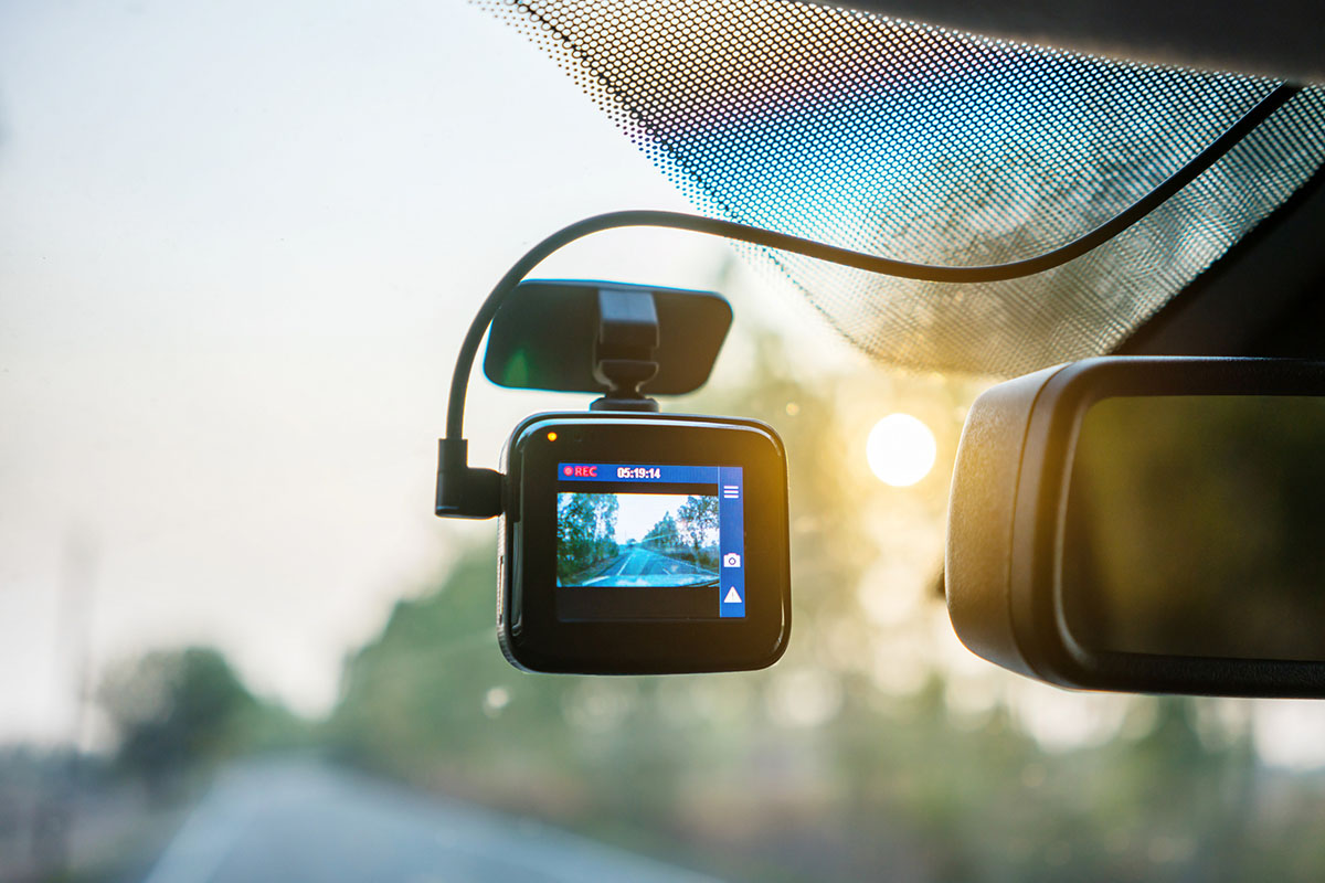Dash camera mounted on a vehicle windshield beside the rearview mirror, its screen recording the road ahead in soft morning light