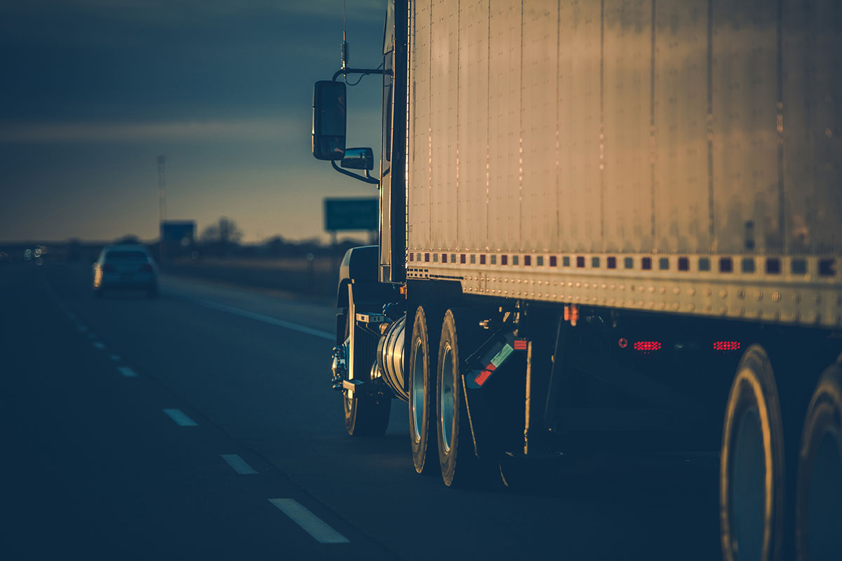 Side view of a semi truck and silver trailer rolling along a highway at dusk, a car ahead in the next lane and road signs in the distance