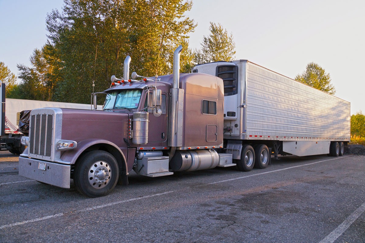 Burgundy long-nose semi truck with a refrigerated trailer parked alone at a truck stop in early morning light, a sunshade across its windshield