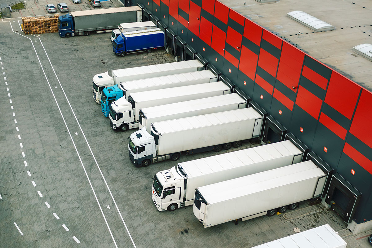 Aerial view of white semi trucks and trailers backed into numbered loading dock doors along a red and charcoal distribution warehouse