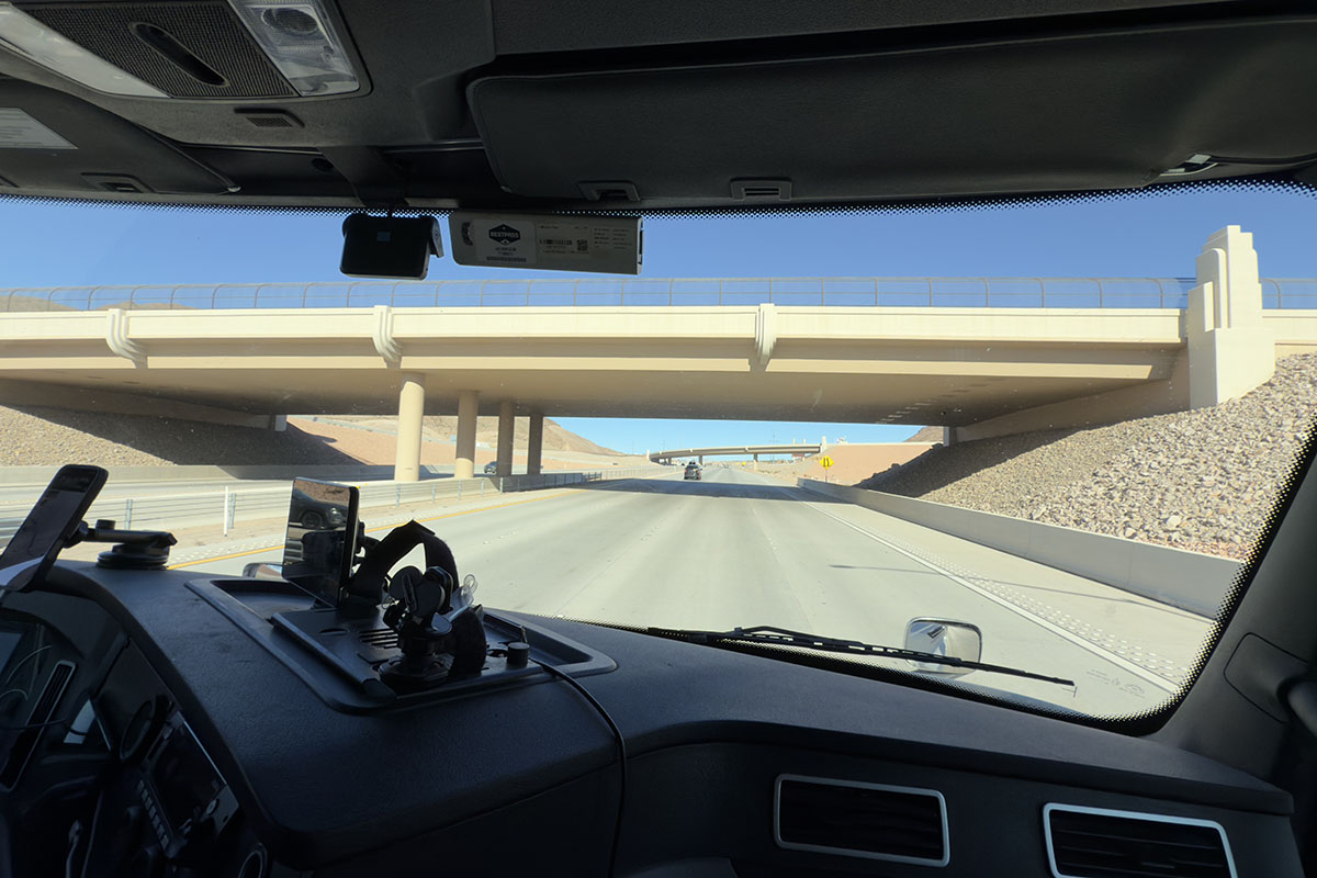 View through the windshield of a truck cab with a dash-mounted camera and toll transponder, approaching a highway overpass under a clear sky