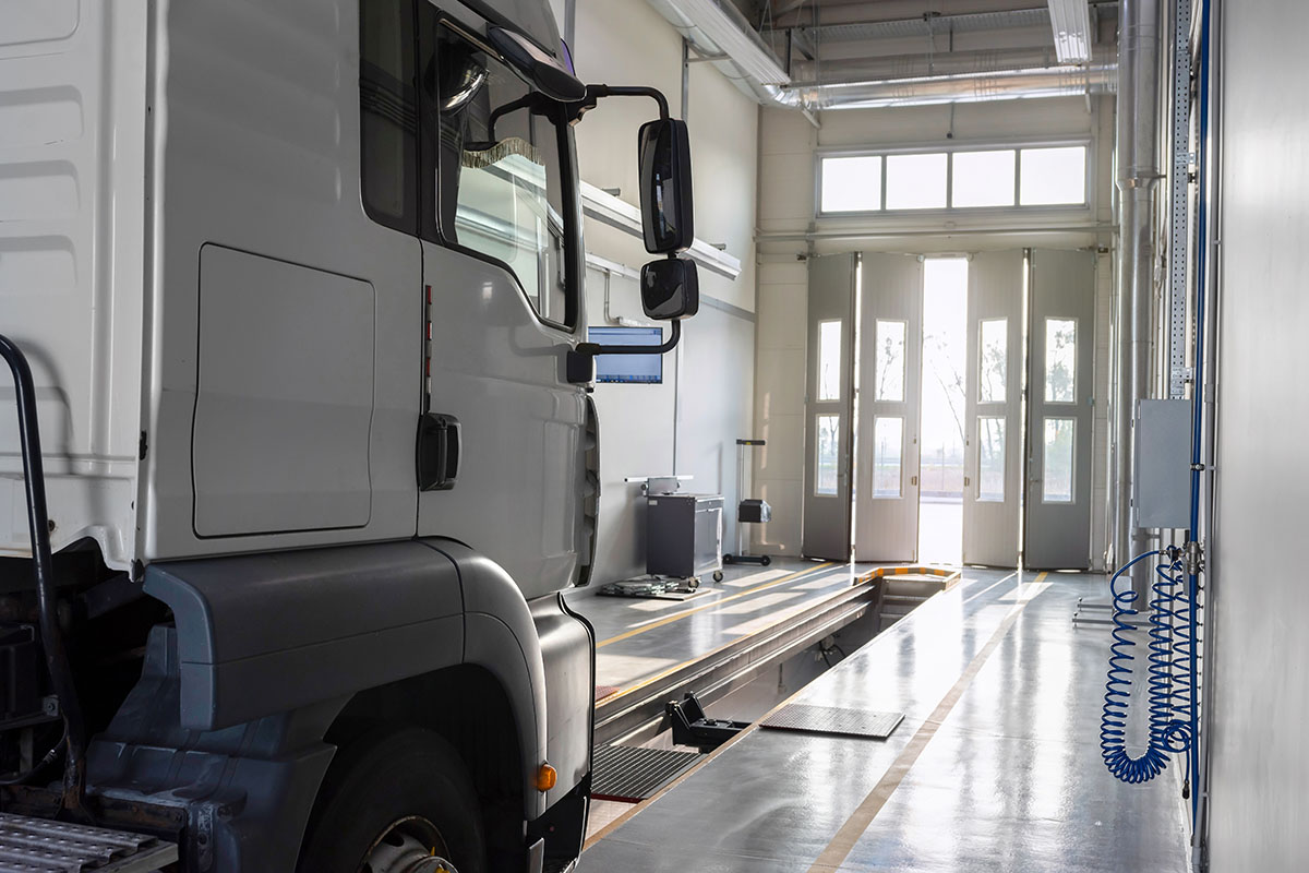White heavy truck cab parked beside an inspection pit in a clean, brightly lit truck service bay, its open doors letting in daylight at the far end