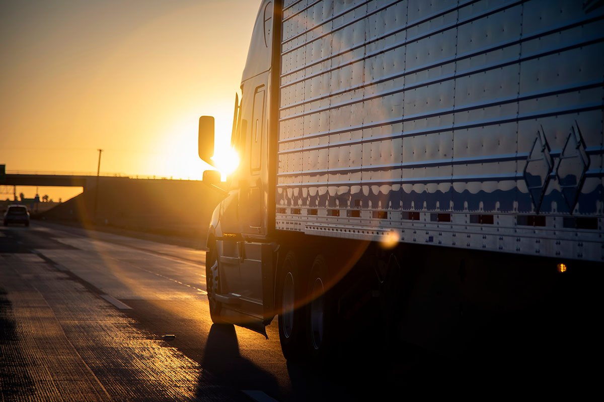 Long-haul semi truck with a silver trailer on a highway at sunrise, the low sun flaring behind its cab and an overpass in the distance