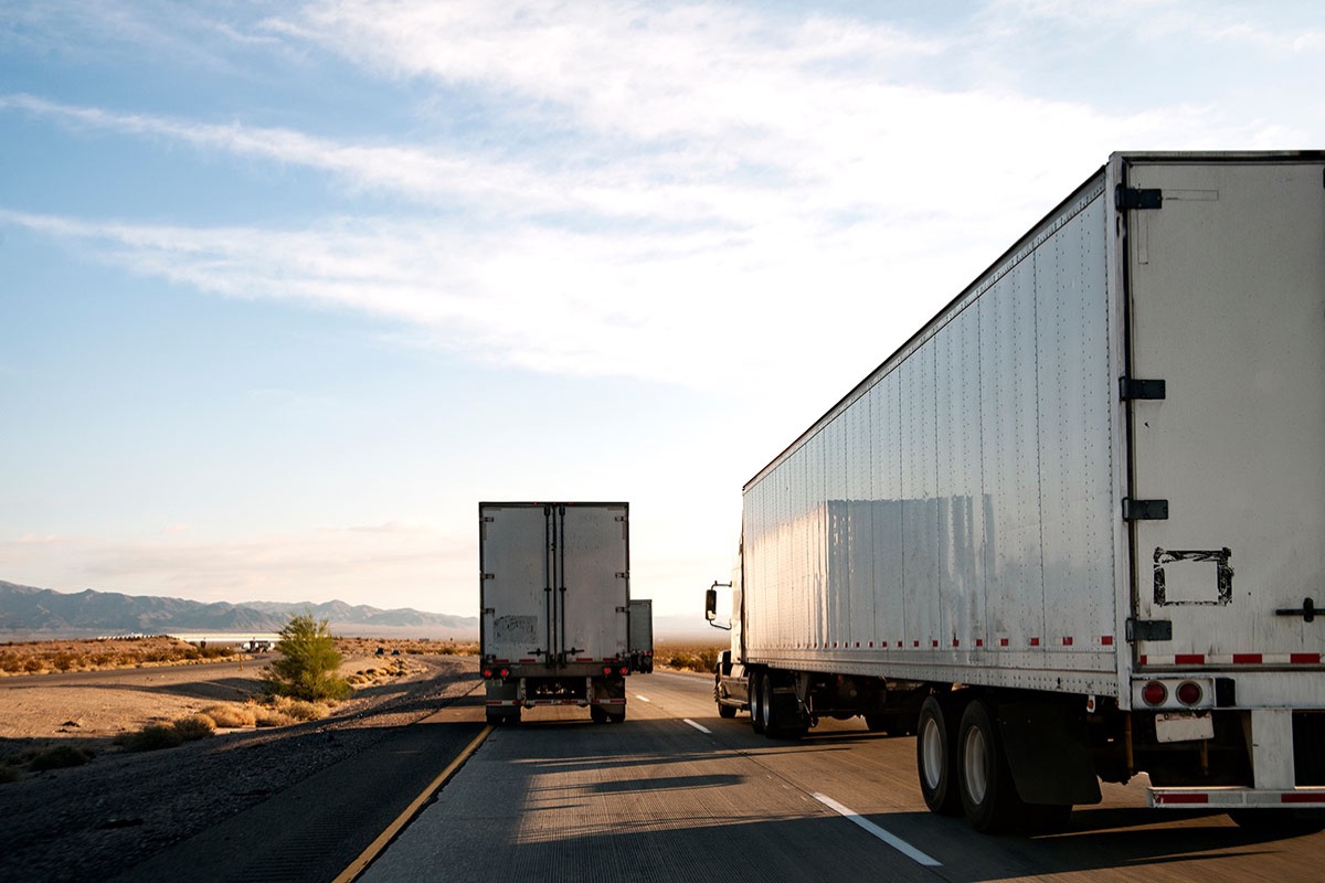 Two white semi trailers heading down a desert interstate toward distant mountains under a pale evening sky