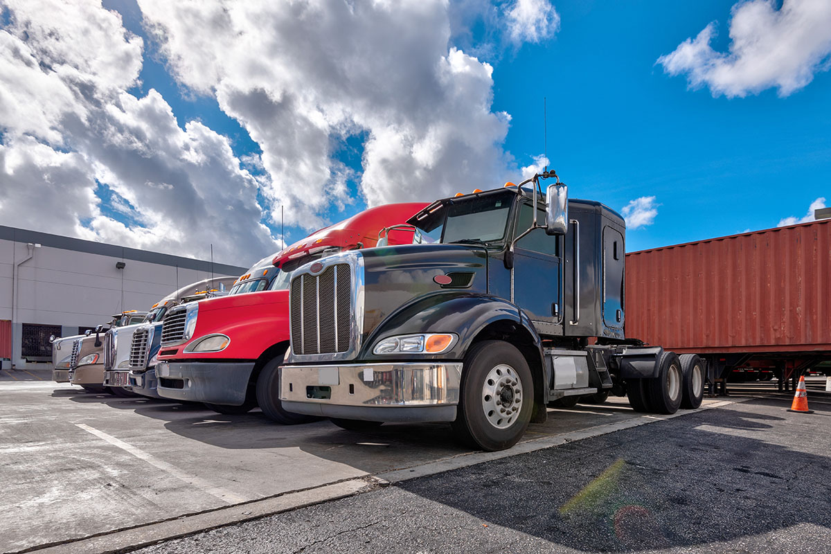 Row of semi truck tractors in black, red and white parked nose to nose outside a logistics warehouse beside a red shipping container under a cloudy blue sky