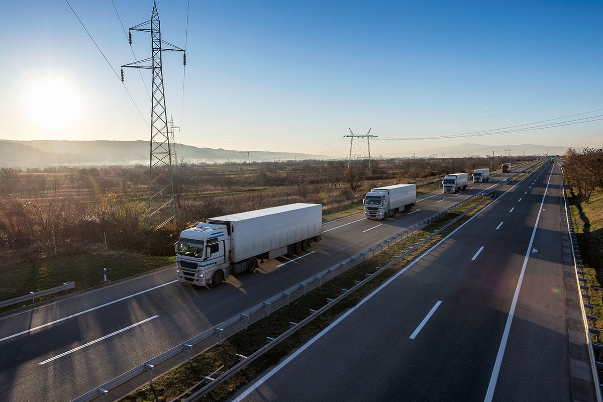 Convoy of white semi trucks travelling in single file along a divided highway past power pylons and open farmland under a low sun