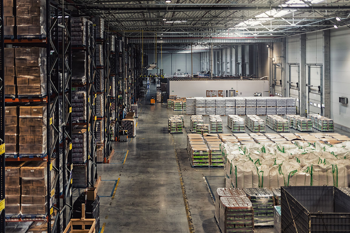 Overhead view of a large distribution warehouse with tall racking of boxed stock on the left and rows of shrink-wrapped pallets and bulk bags covering the floor