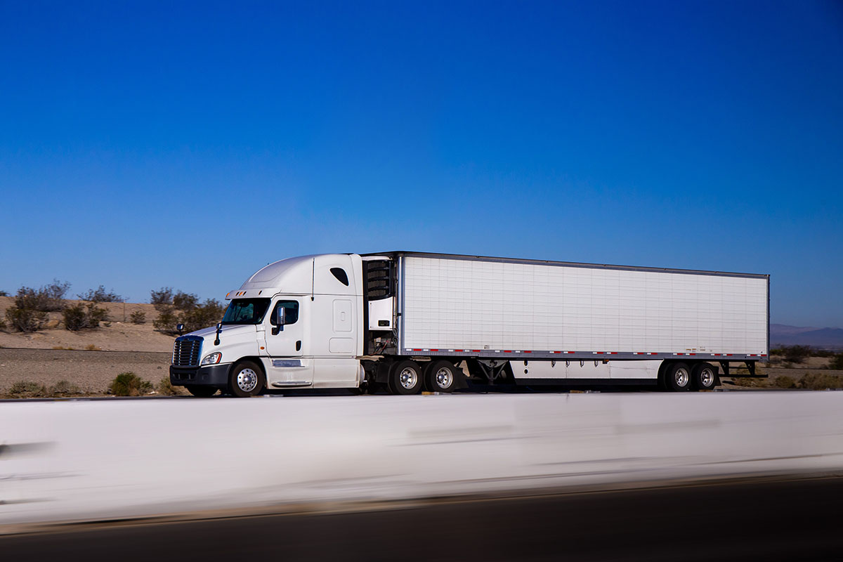 White semi truck with a refrigerated trailer driving along a desert highway under a clear blue sky