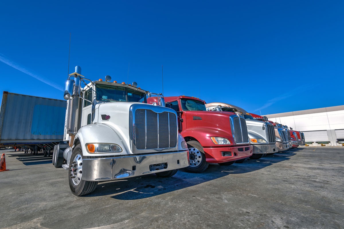 Row of white, red and silver semi truck tractors parked side by side at a distribution center lot under a deep blue sky