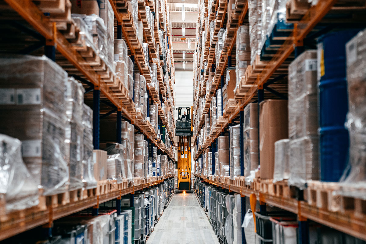 Long high-bay warehouse aisle lined with orange racking full of shrink-wrapped pallets, with a yellow order picker at the far end