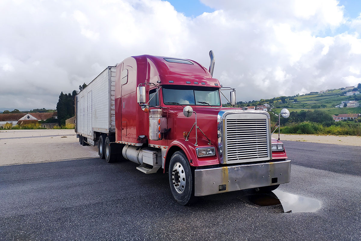 Red long-nose semi truck with a white box trailer parked on an open asphalt lot under a cloudy sky, a rain puddle beside its front bumper