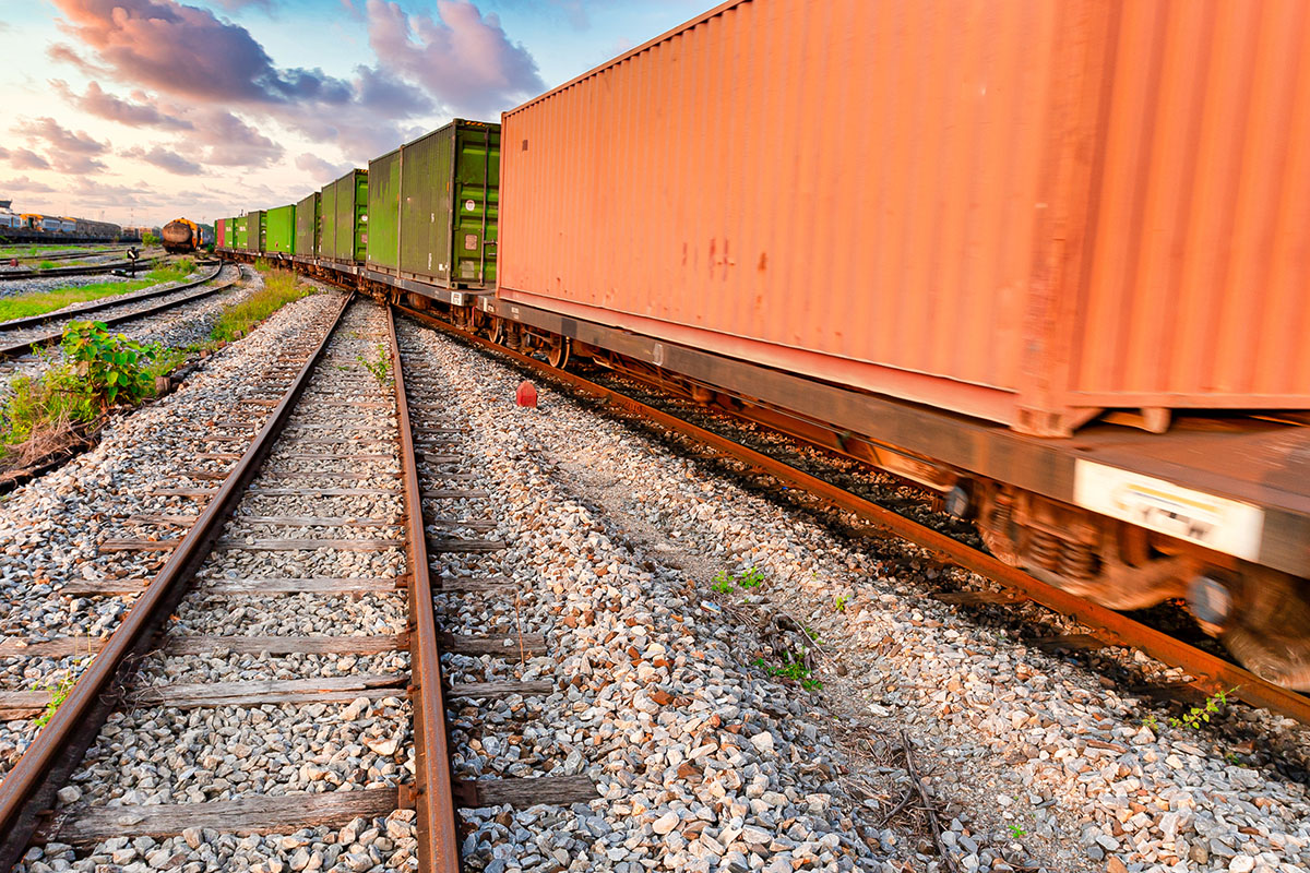 Freight train loaded with orange and green shipping containers rolling past empty rail tracks and gravel ballast under a pink sunset sky