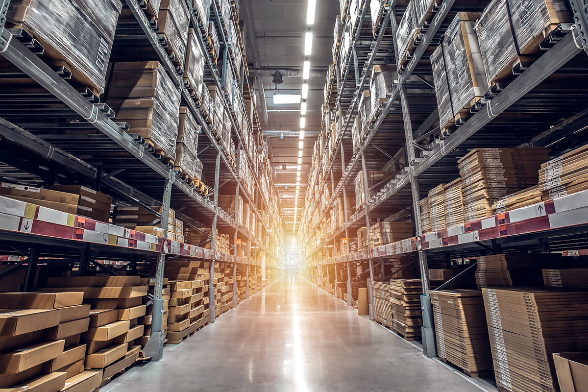 Long warehouse aisle lined with tall steel racking packed with shrink-wrapped pallets and flat cardboard cartons